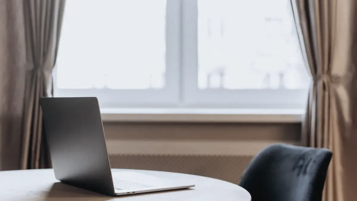 A laptop on a round wooden table by a bright window